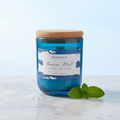 Unlit House of Wick Ocean Mint 12 oz blue glass candle with a cork lid, styled on a white marble surface next to fresh mint leaves and sea salt crystals.
