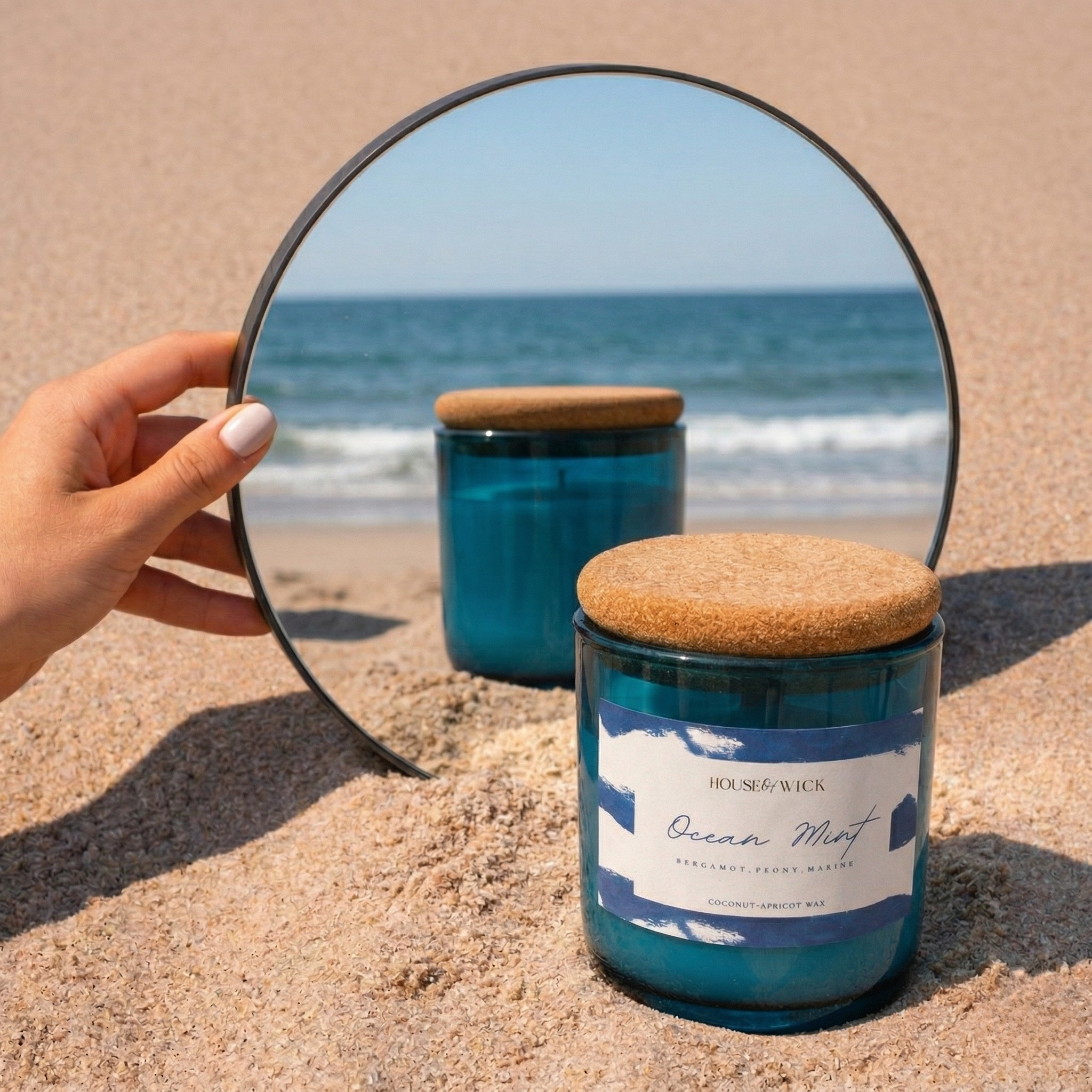 House of Wick Ocean Mint 12 oz coconut-apricot wax candle in a blue glass jar with a cork lid, reflecting in a round mirror placed on sunlit beach sand.
