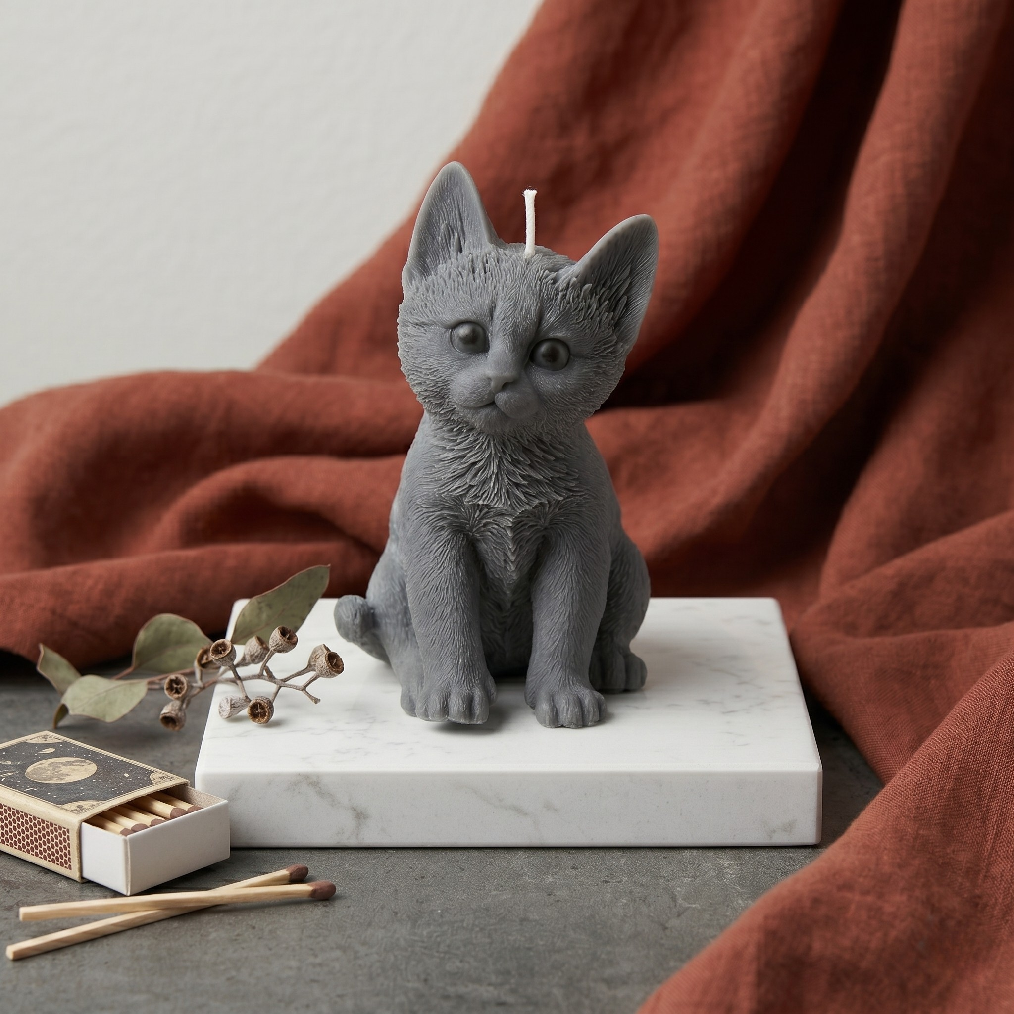 Grey cat-shaped decorative candle on a marble base, styled against a warm terracotta fabric backdrop with vintage matches.