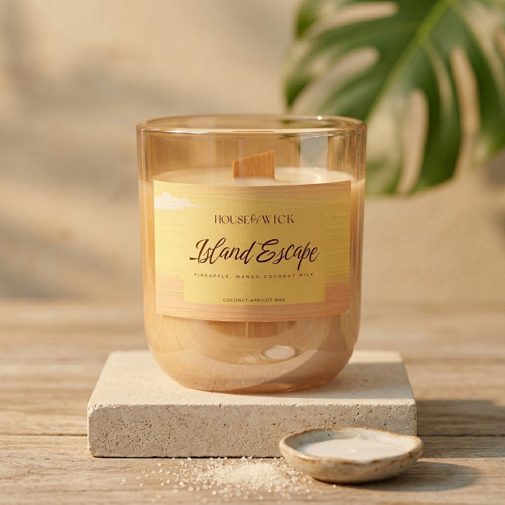 Open House of Wick Island Escape 12 oz glass jar candle displaying a wooden wick, styled on a textured stone block with scattered sugar and a small dish of coconut milk, featuring a monstera leaf in the background.