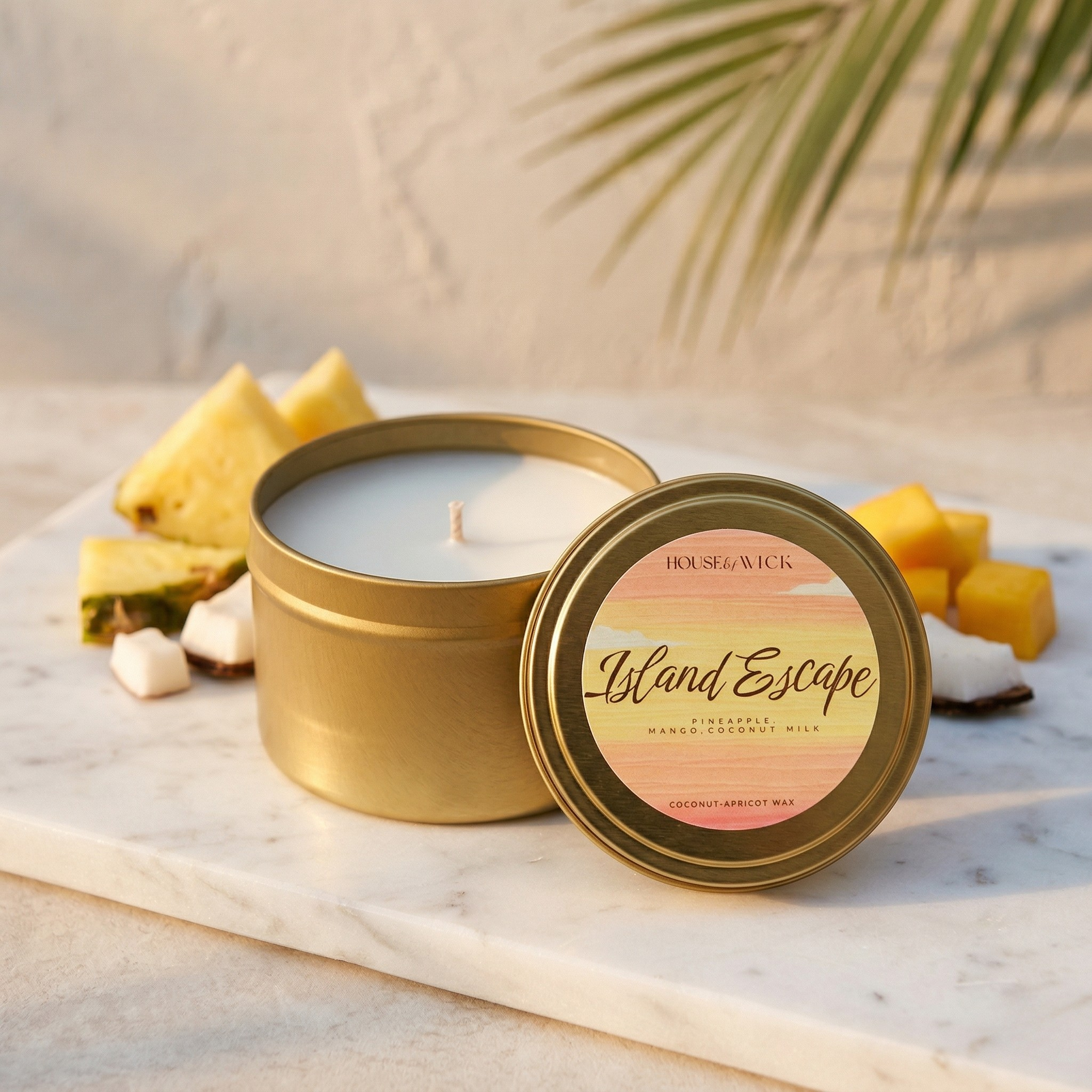 Open House of Wick Island Escape 6 oz gold tin travel candle displaying white wax, leaning against its lid on a white marble board. It is surrounded by fresh pineapple, mango, and coconut chunks.