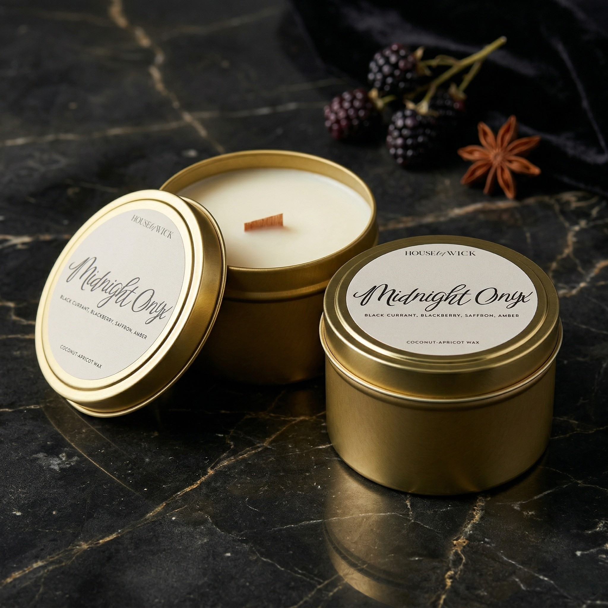 Two House of Wick Midnight Onyx 6 oz gold tin candles—one open showing the wooden wick and one closed—displayed on dark marble with fresh blackberries and star anise.
