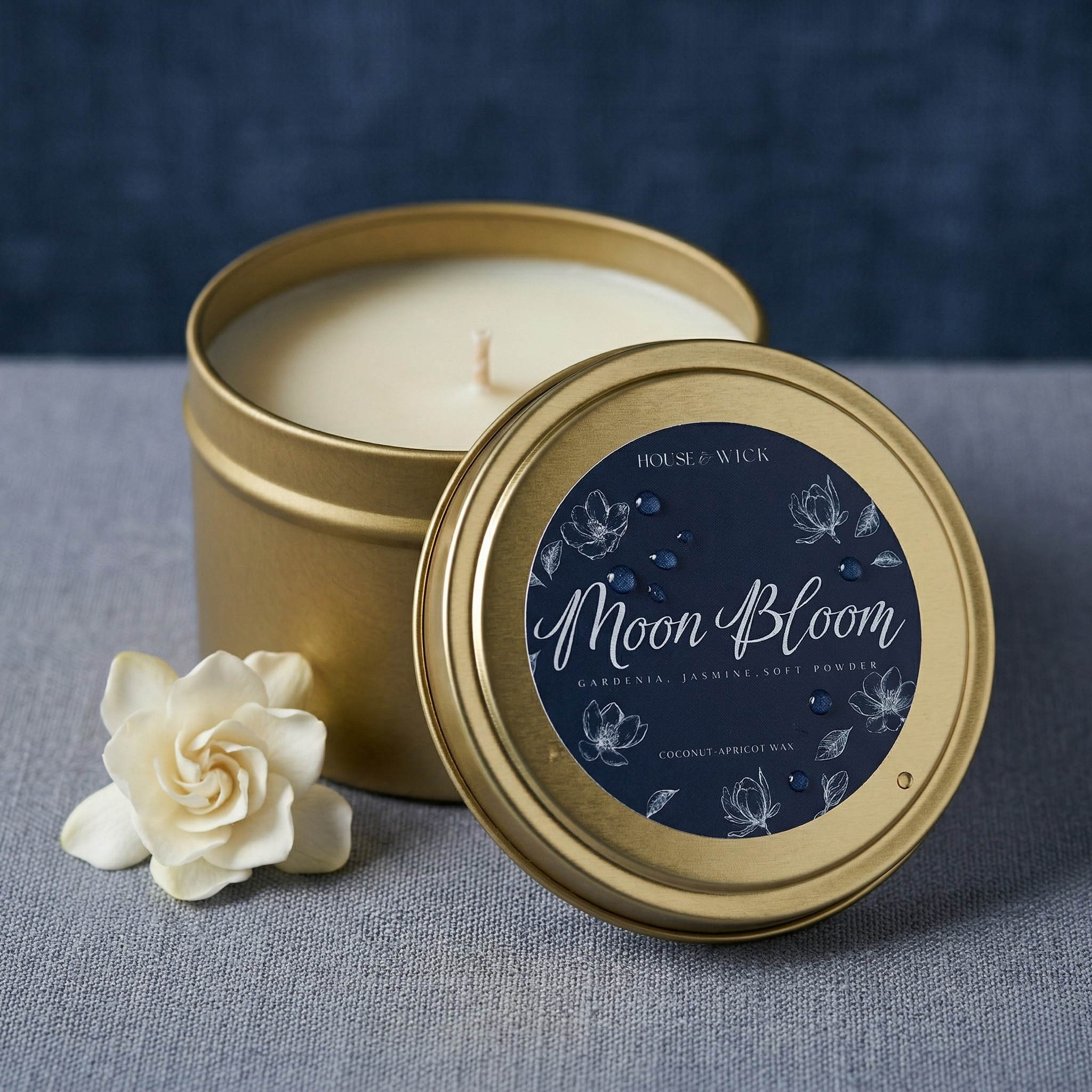 Open House of Wick Moon Bloom 6 oz gold tin travel candle displaying white wax, leaning against its blue floral label lid, styled with a fresh white gardenia flower on a textured grey surface.