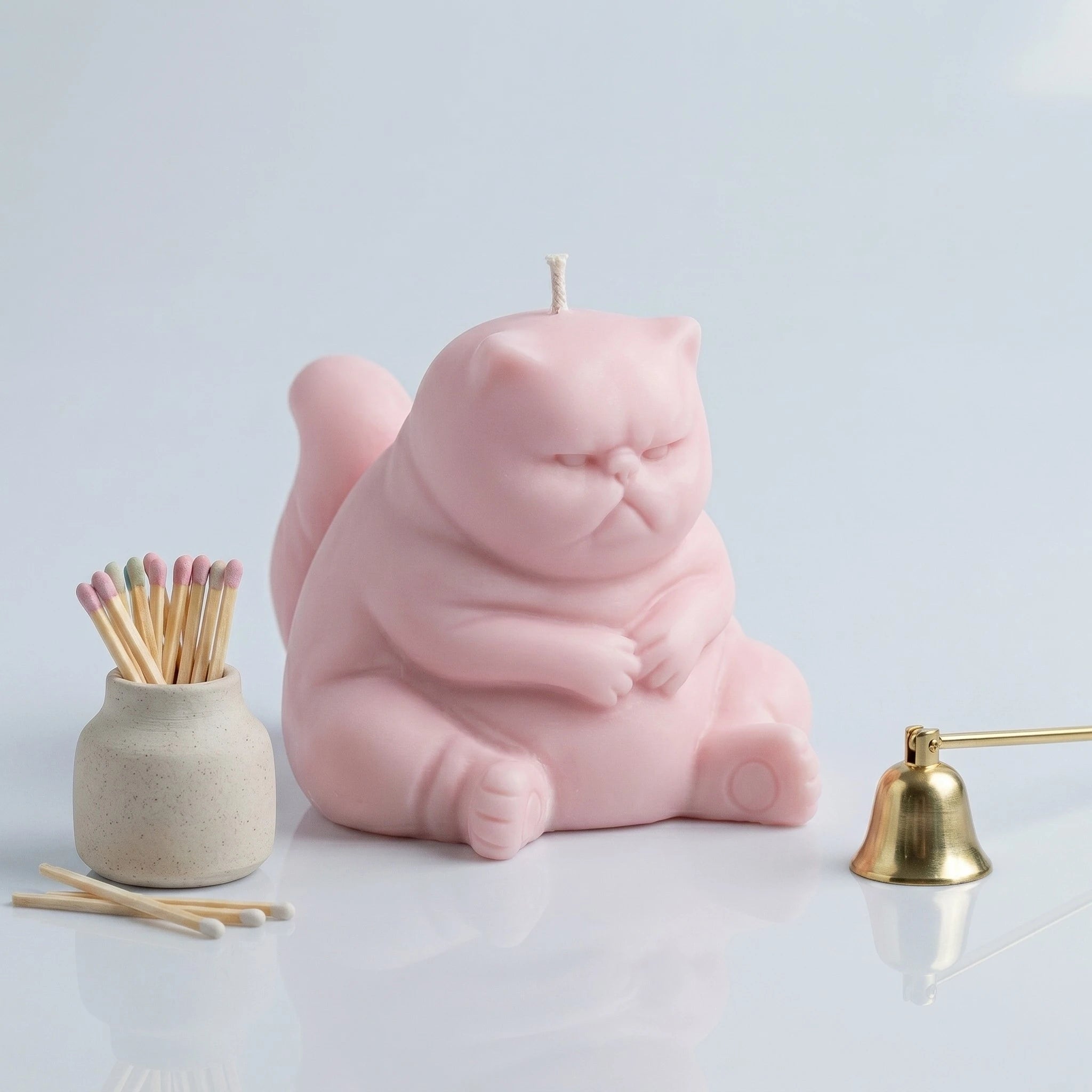 Pastel pink chubby cat candle with folded paws on clean background with match jar and gold bell