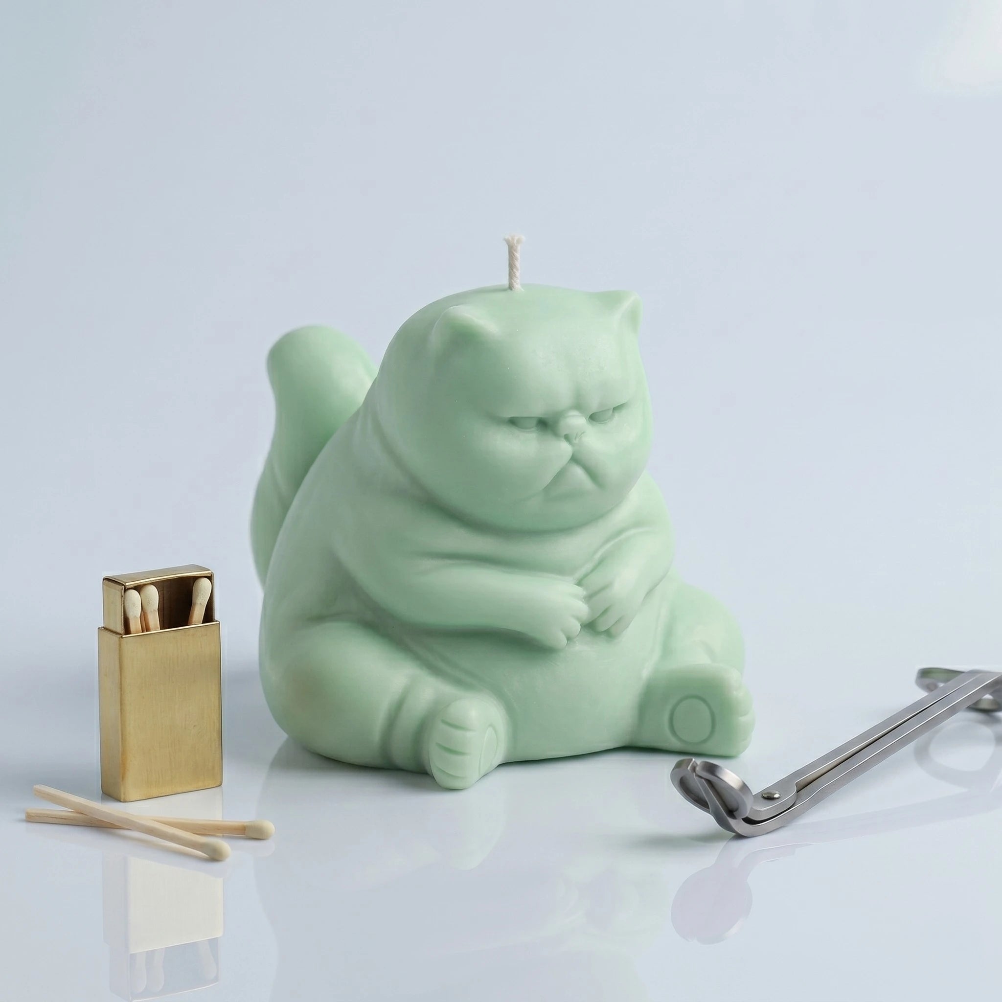 Pastel sage green chubby cat candle with folded paws on clean background with gold lighter and wick trimmer