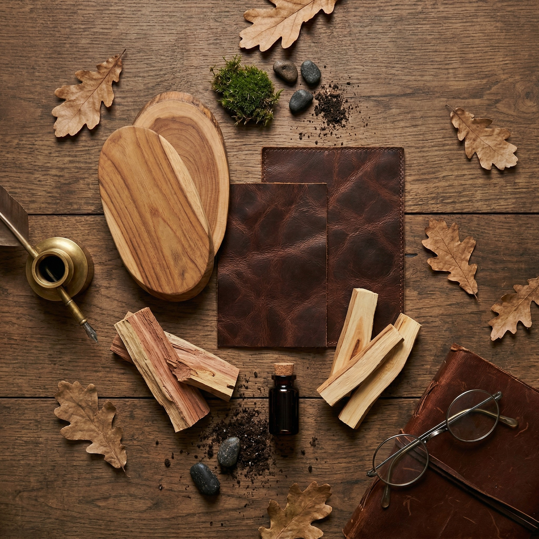 Flat lay of House of Wick Vintage Library fragrance scent notes, featuring smooth teakwood, dark leather swatches, cedarwood sticks, an amber fragrance bottle, and oak leaves on a rustic wooden table.