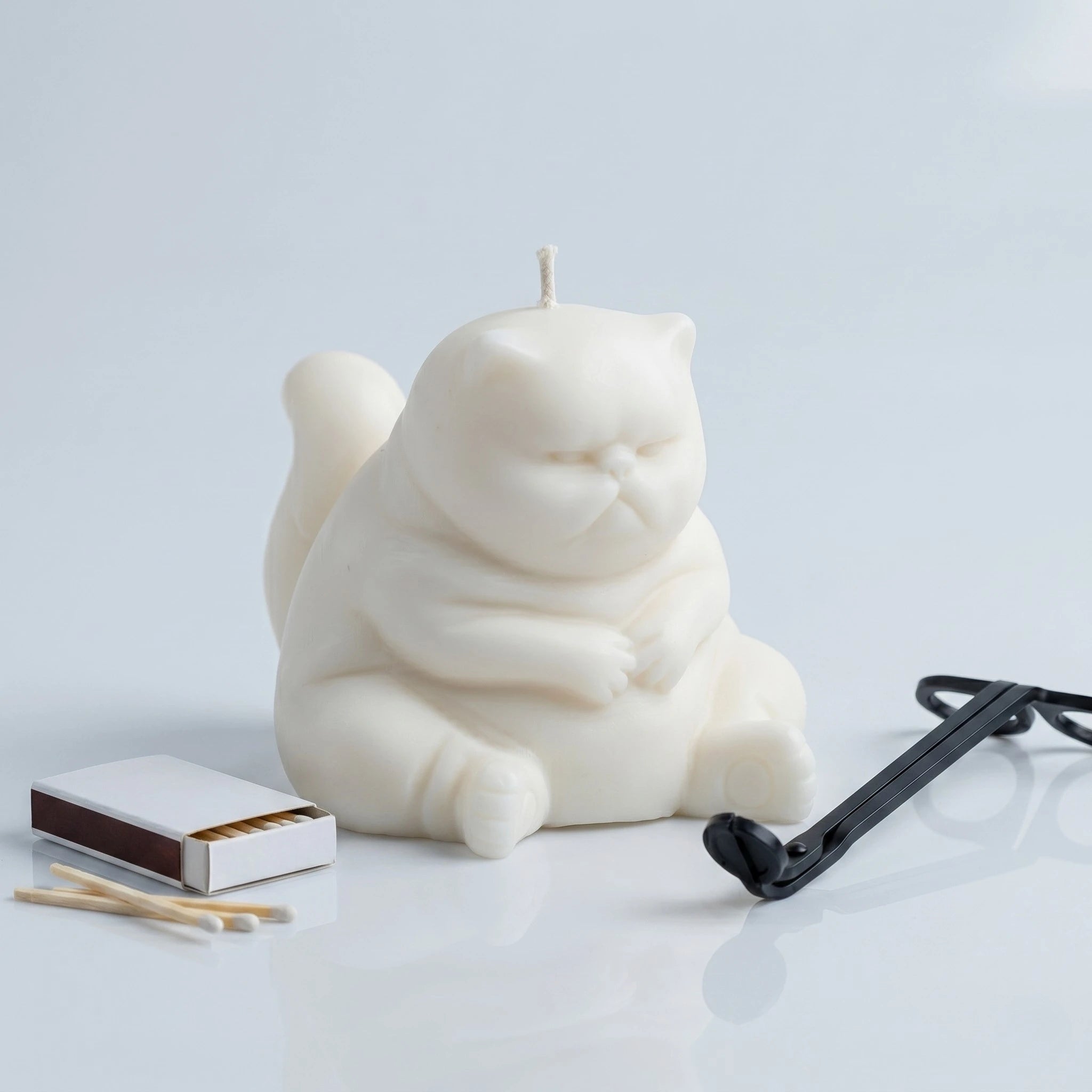 White chubby cat candle with folded paws on clean background with matchbox and wick trimmer
