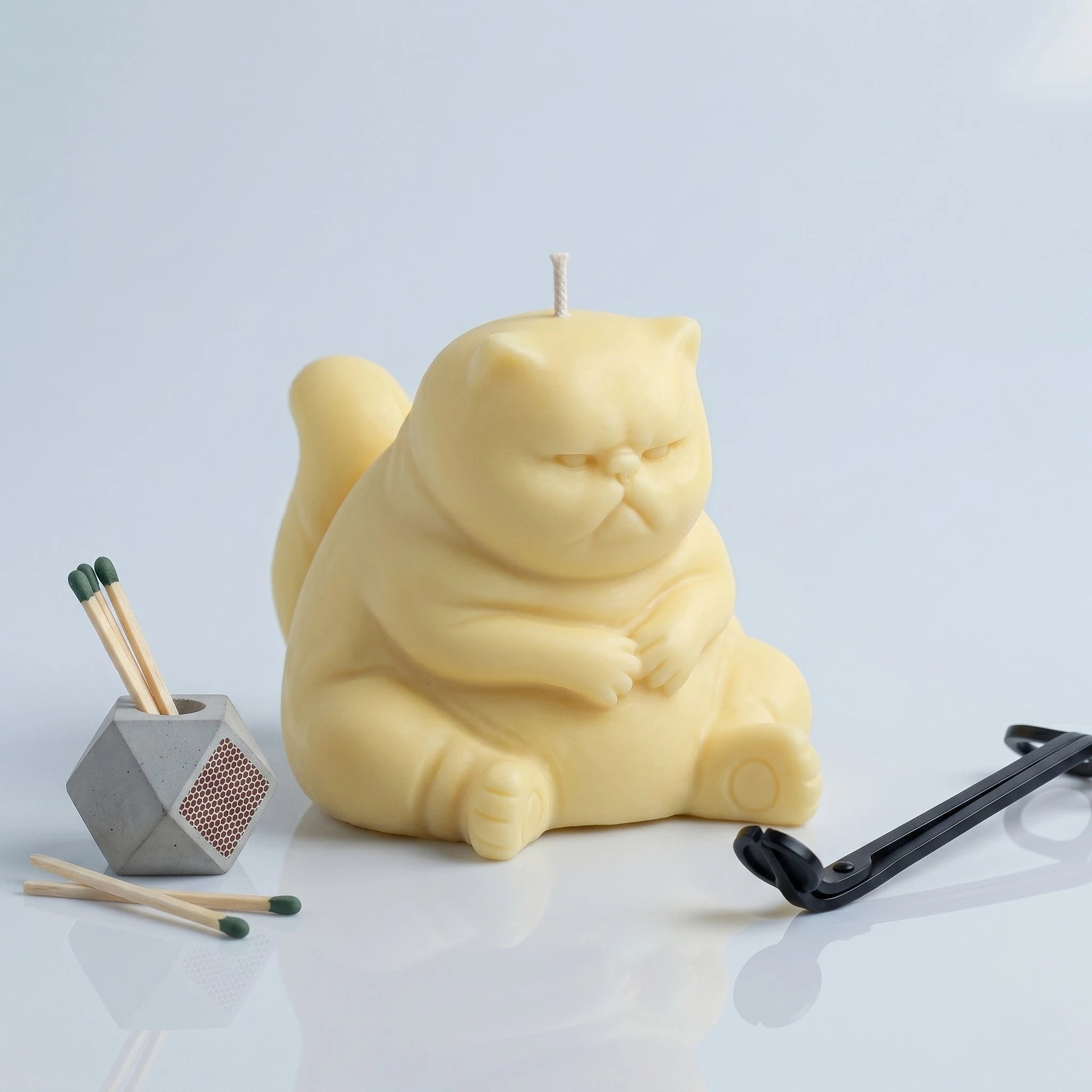 Pastel yellow chubby cat candle with folded paws on clean background with match holder and wick trimmer